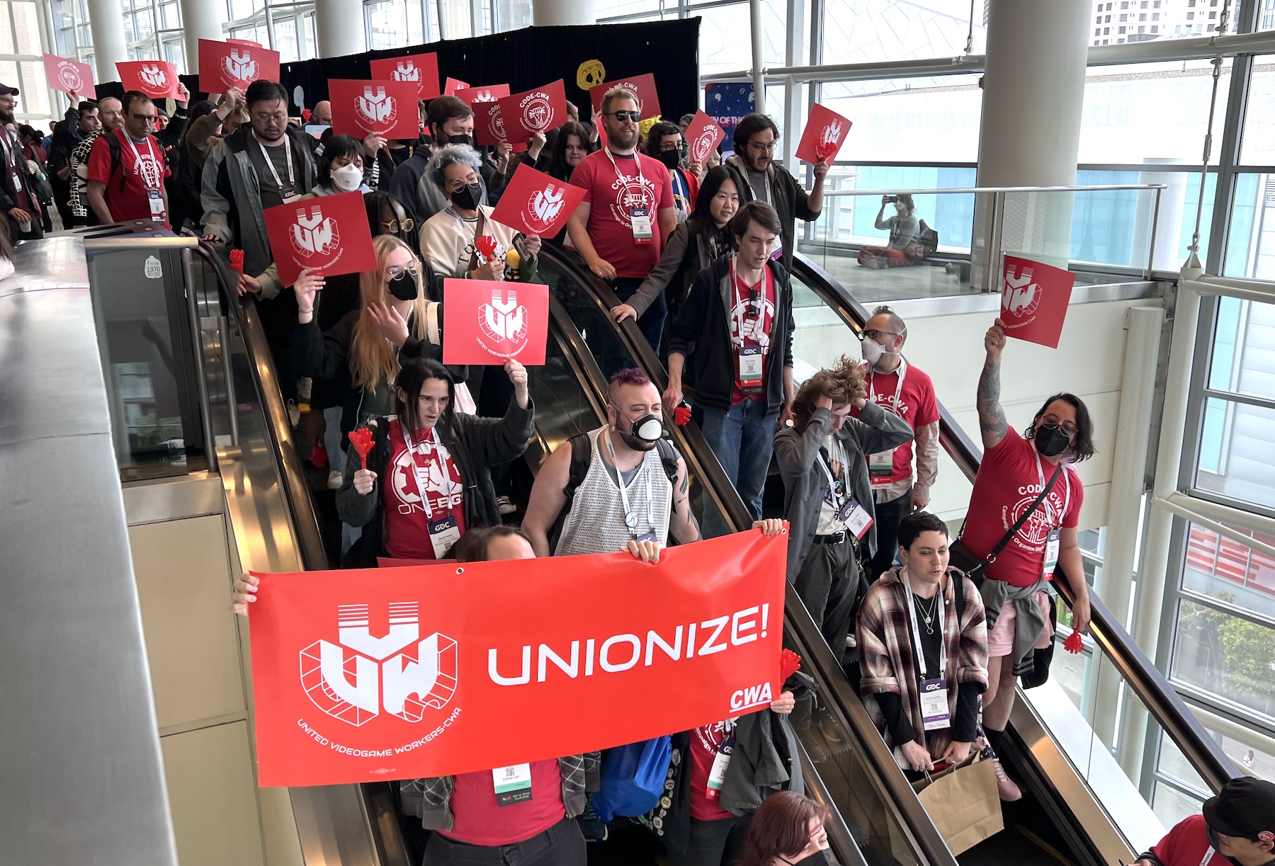 Workers wearing GDC conference passes fill an escalator going down. Many hold signs that say UVW and one holds a larger banner that says UVW: UNIONIZE!